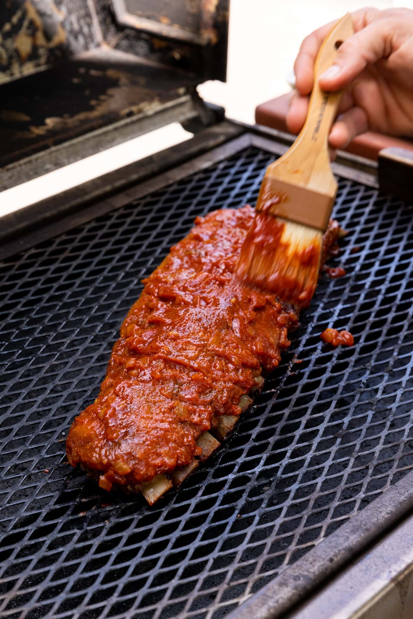 Hand holding brush brushing sweet barbecue sauce on top of ribs cooking on the grill top.