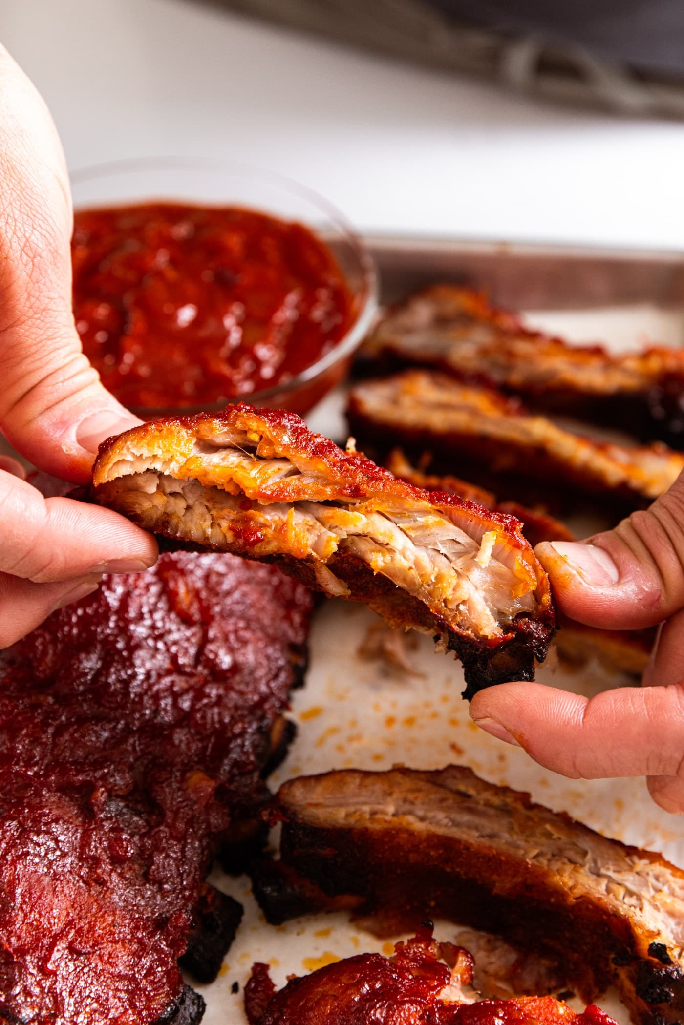 Two hands holding one baby back rib with barbecue sauce slathered all over with remaining ribs in background.