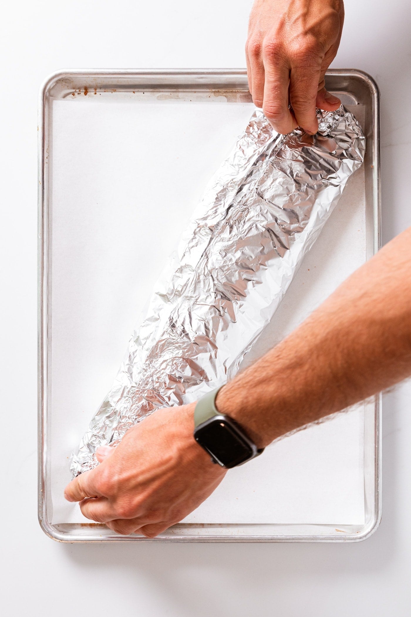 Two hands crimping edges of tin foil together at opposite ends of foil with package sitting on baking dish.