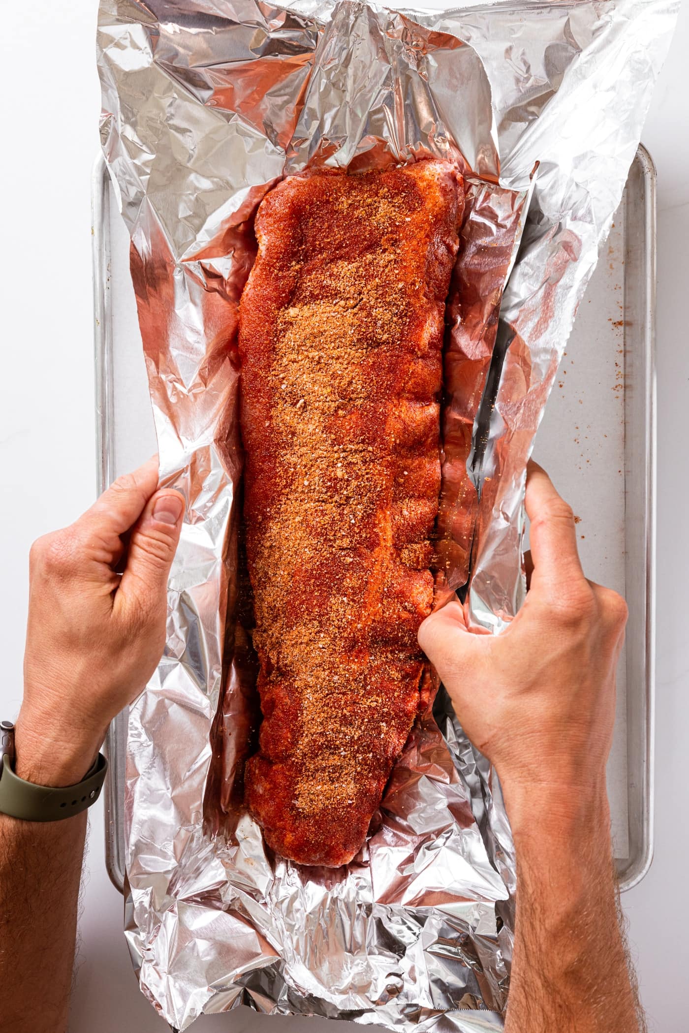 Two hand folding up sides of tin foil with baby back ribs inside.