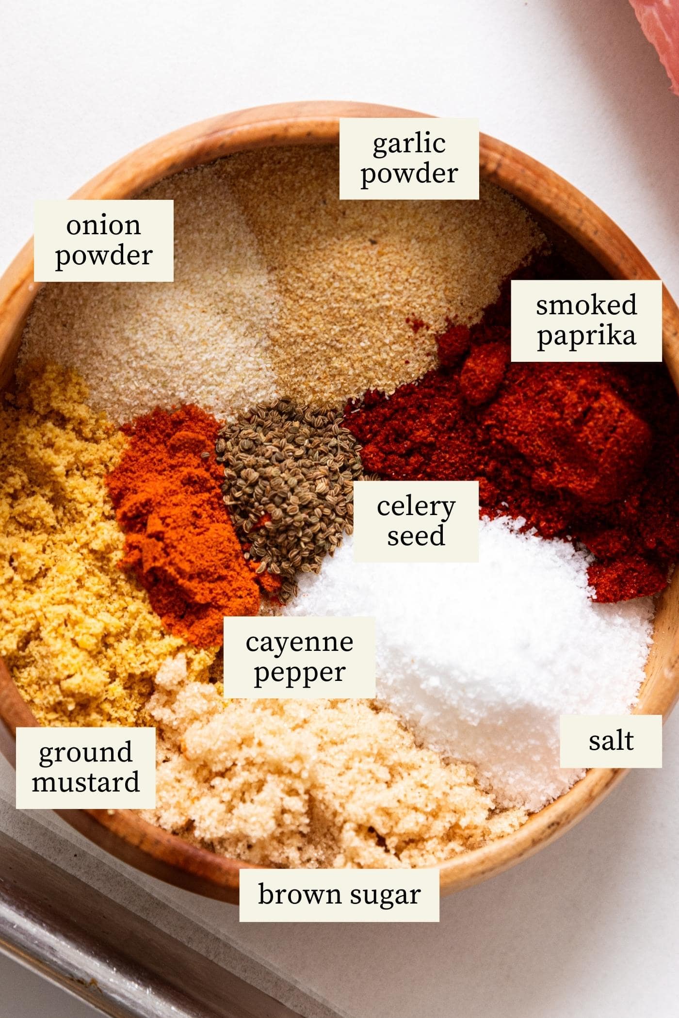 Wooden bowl filled with lots of different spices with words written on top to indicate which spice is which all on white surface.