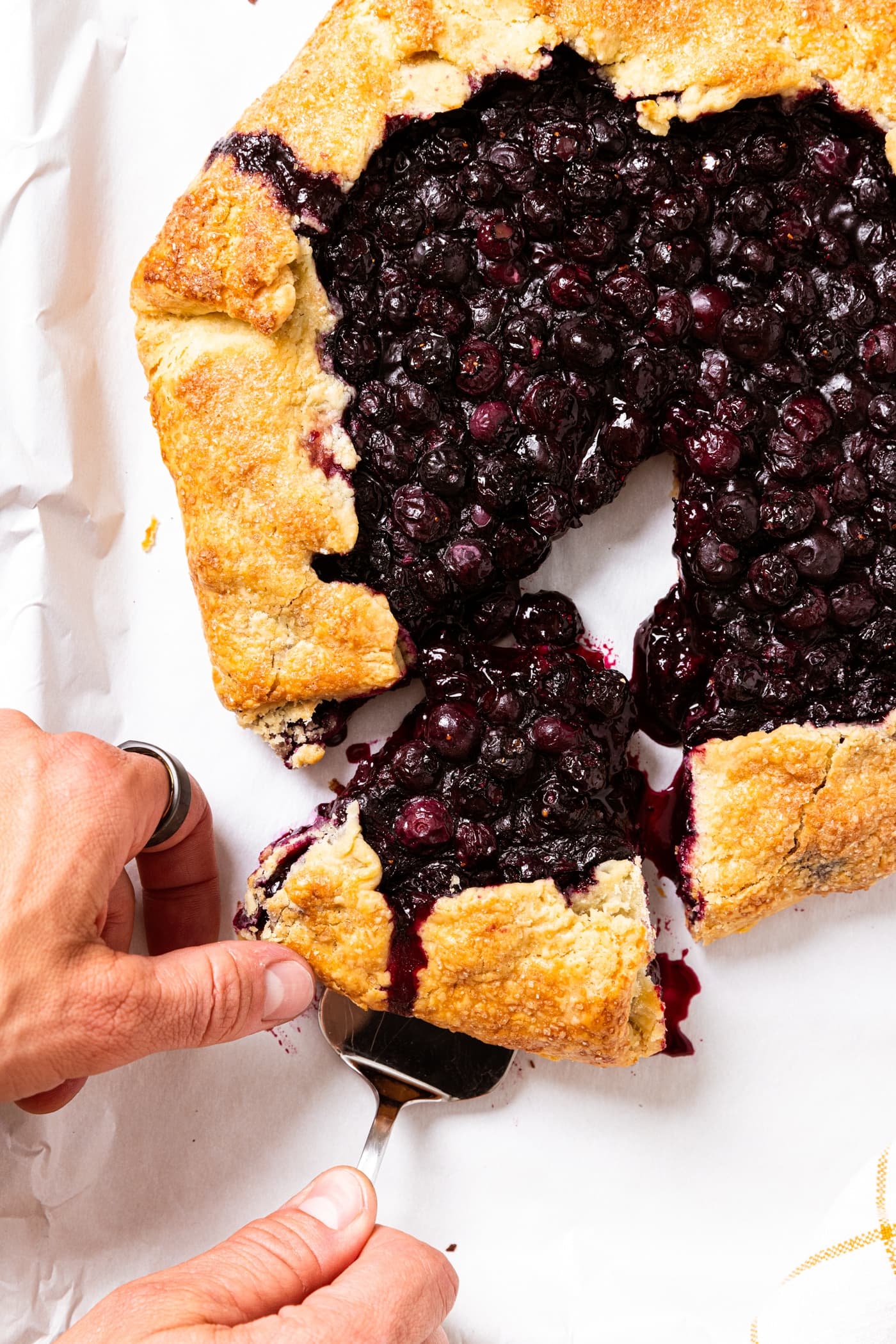 Hand holding pie scoop pulling piece of blueberry galette from rest of galette with white piece of parchment paper underneath.