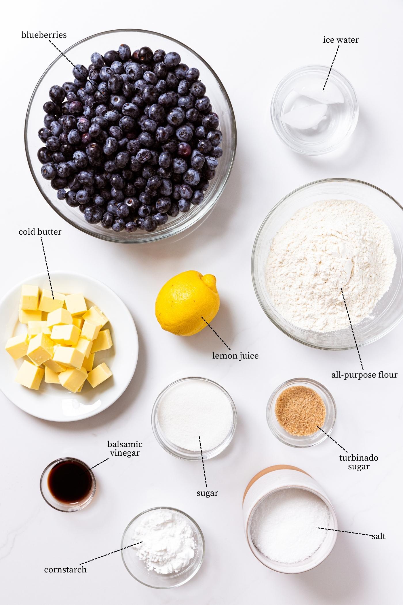 White countertop with ingredients needed to make a blueberry galette including blueberries, lemon, sugar, flour, and butter.