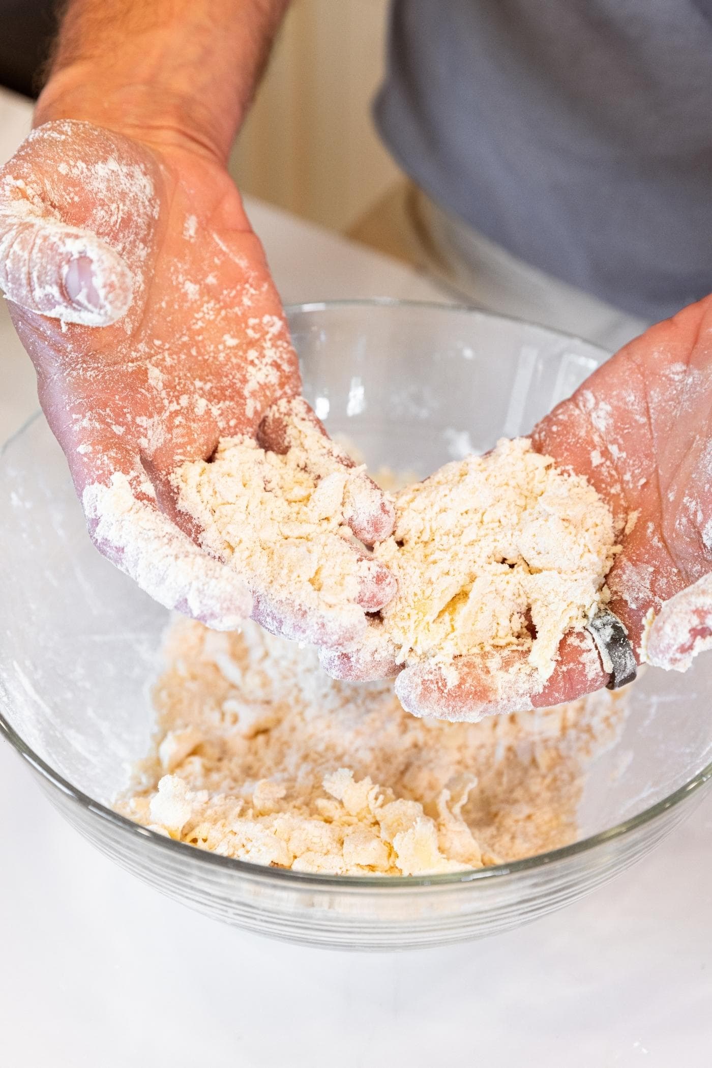 Two hands holding crumbly dough with rest of dough in glass bowl below all with white countertop.