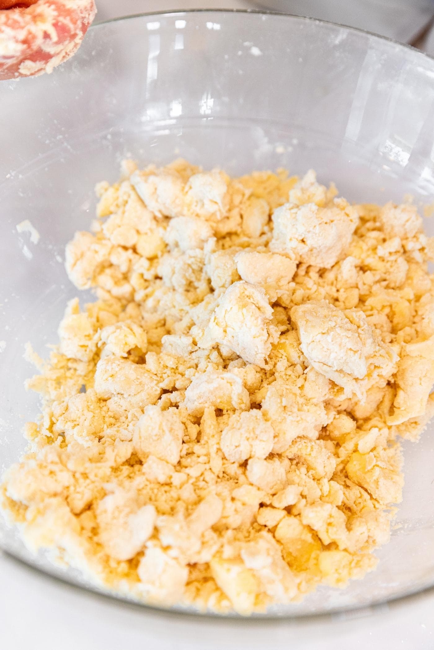 Glass bowl filled with craggy pieces of dough all on white surface with dirty floured hands to the side.