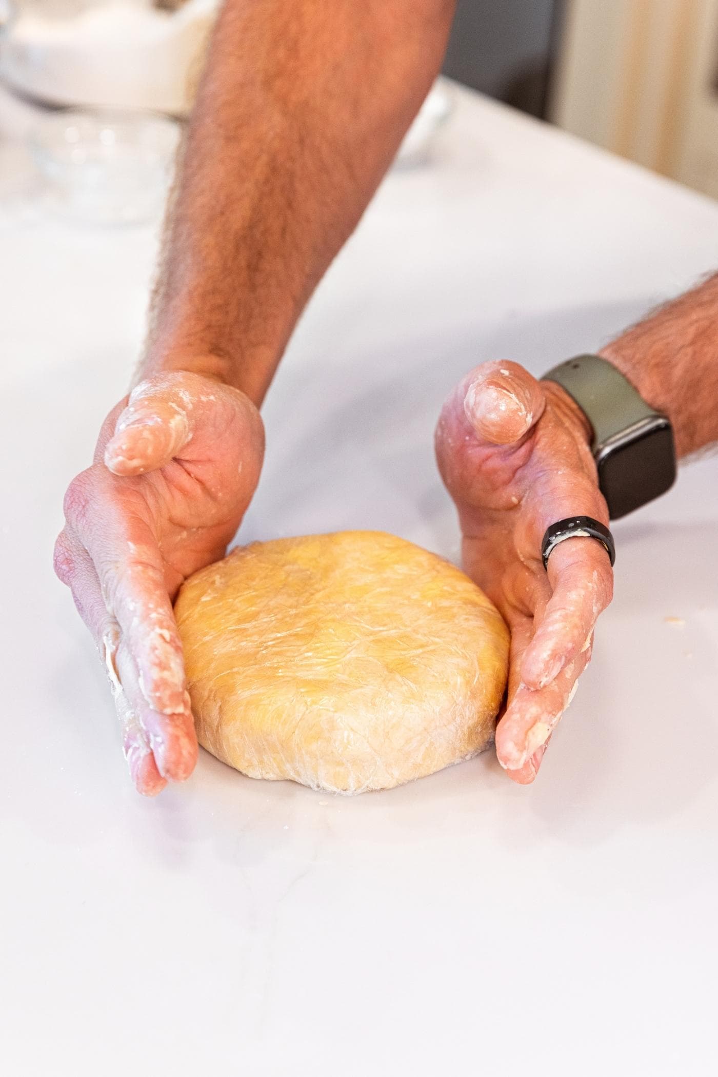 Two hands forming dough in plastic wrap into a circle on white countertop.