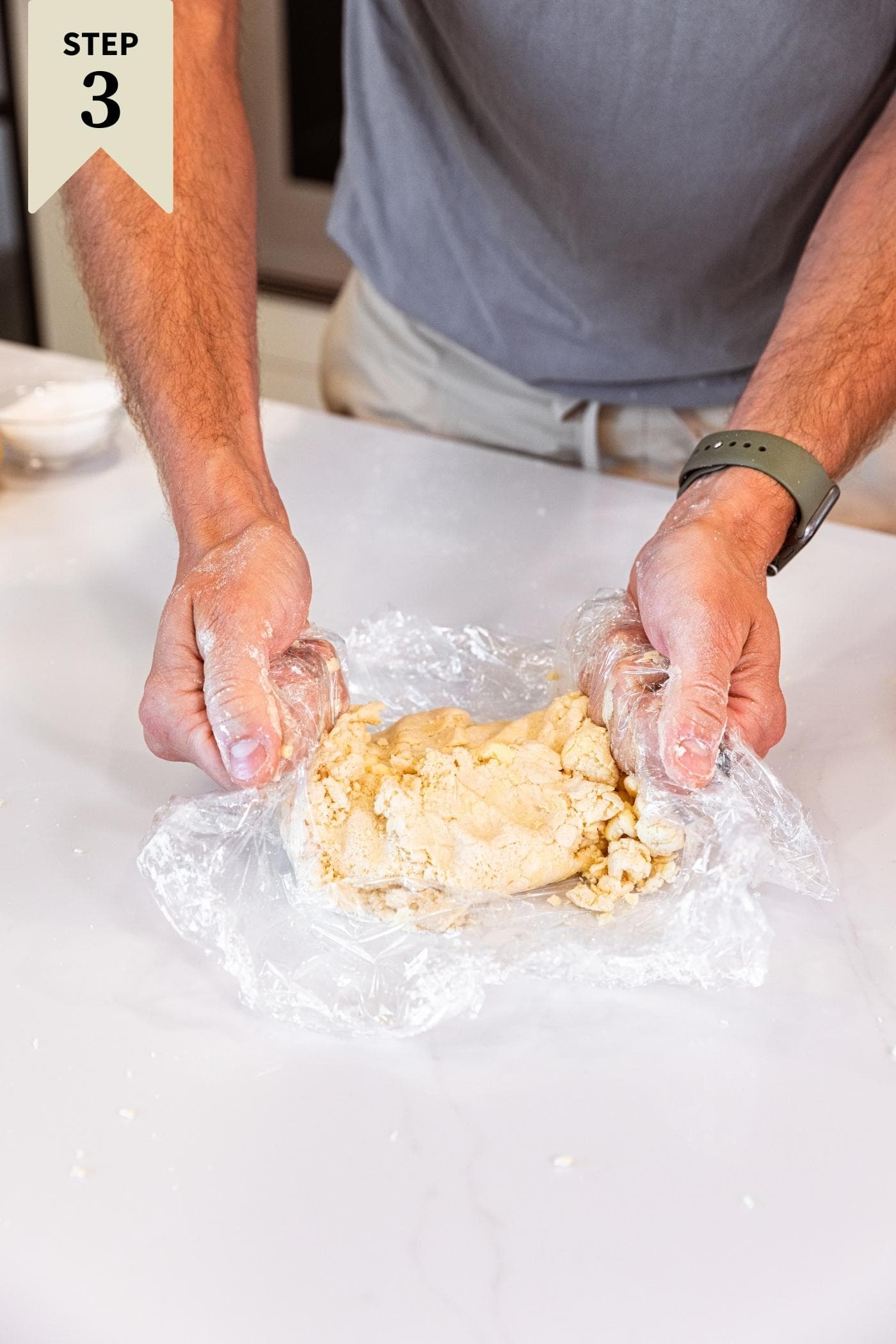 Two hands holding plastic wrap with dough in the middle pushing in from the sides all on white countertop.