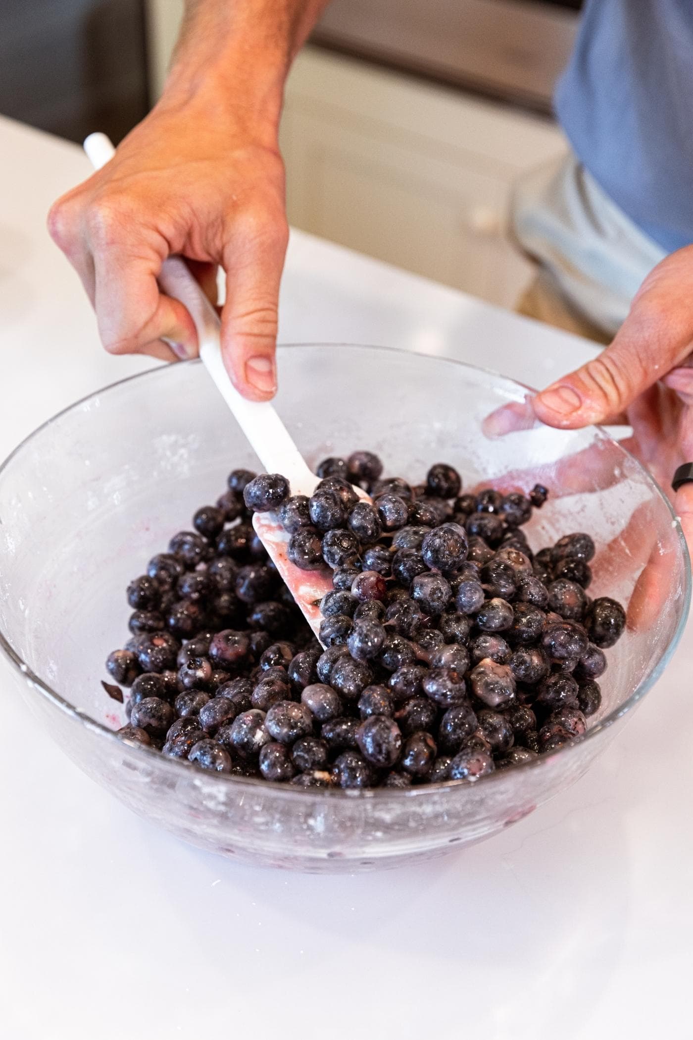 Hand holding white spatula mixing blueberries in glass mixing bowl all on white countertop.