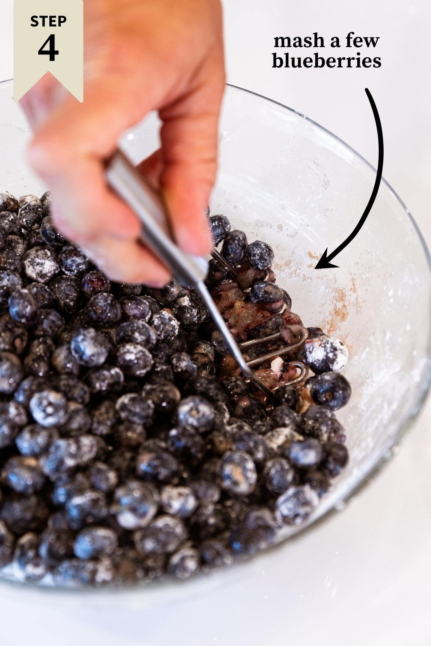 Hand holding potato masher mashing some blueberries in glass bowl.
