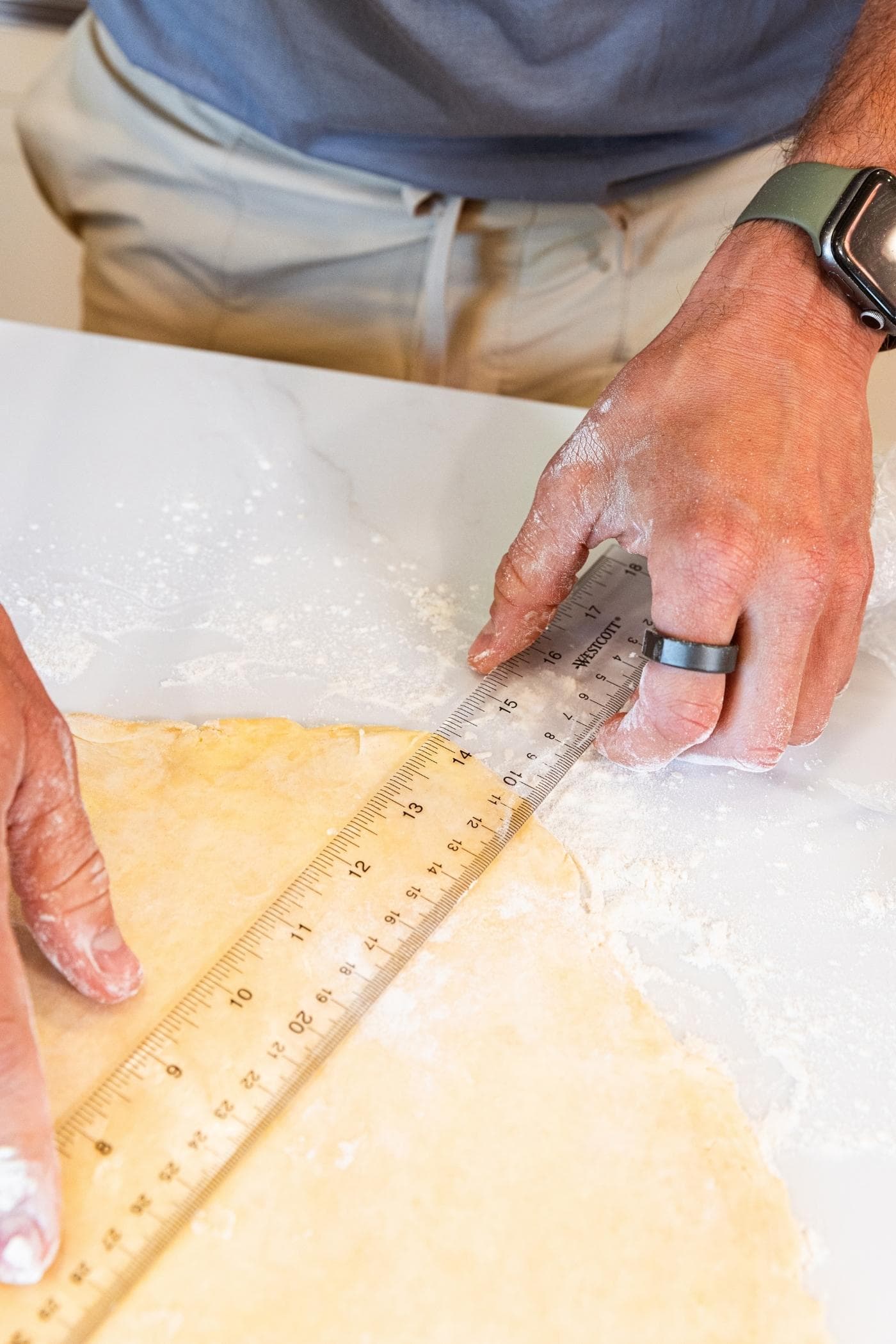 Hand holding ruler showing that a pie crust on white countertop is 14 inches wide.