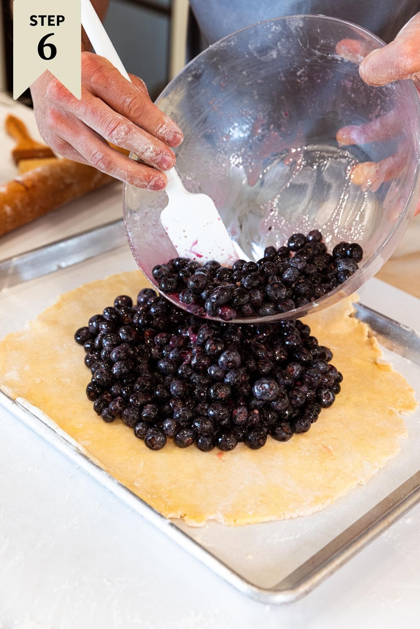 Hand scooping out blueberry filling from glass bowl onto pie crust dough on metal sheet pan.