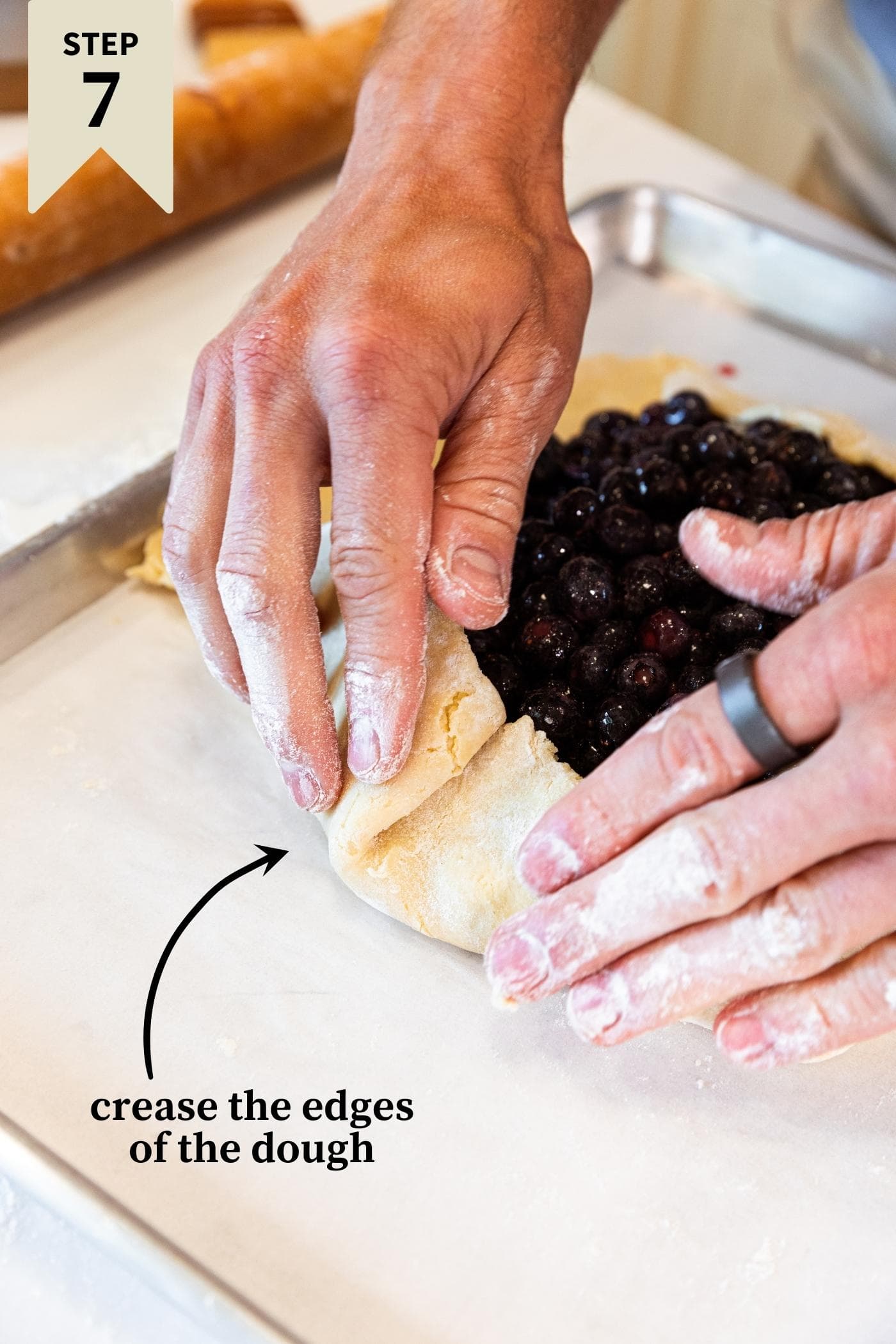 Two hands folding pie crust over itself with blueberry filling in middle of pie all on white countertop.