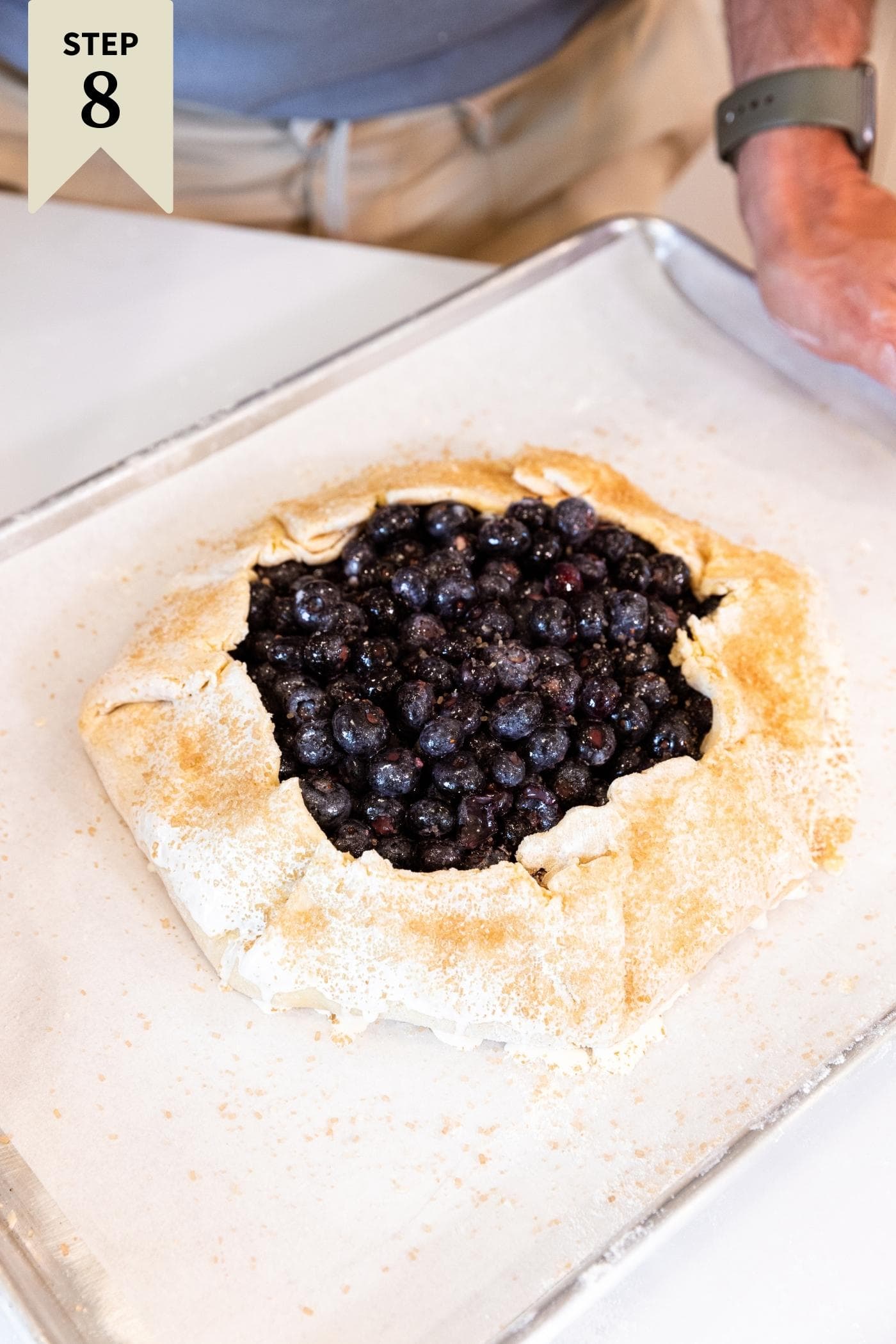 Hand holding baking sheet filled with formed blueberry galette with blueberries inside and unbaked crust surround right before placing in the oven.