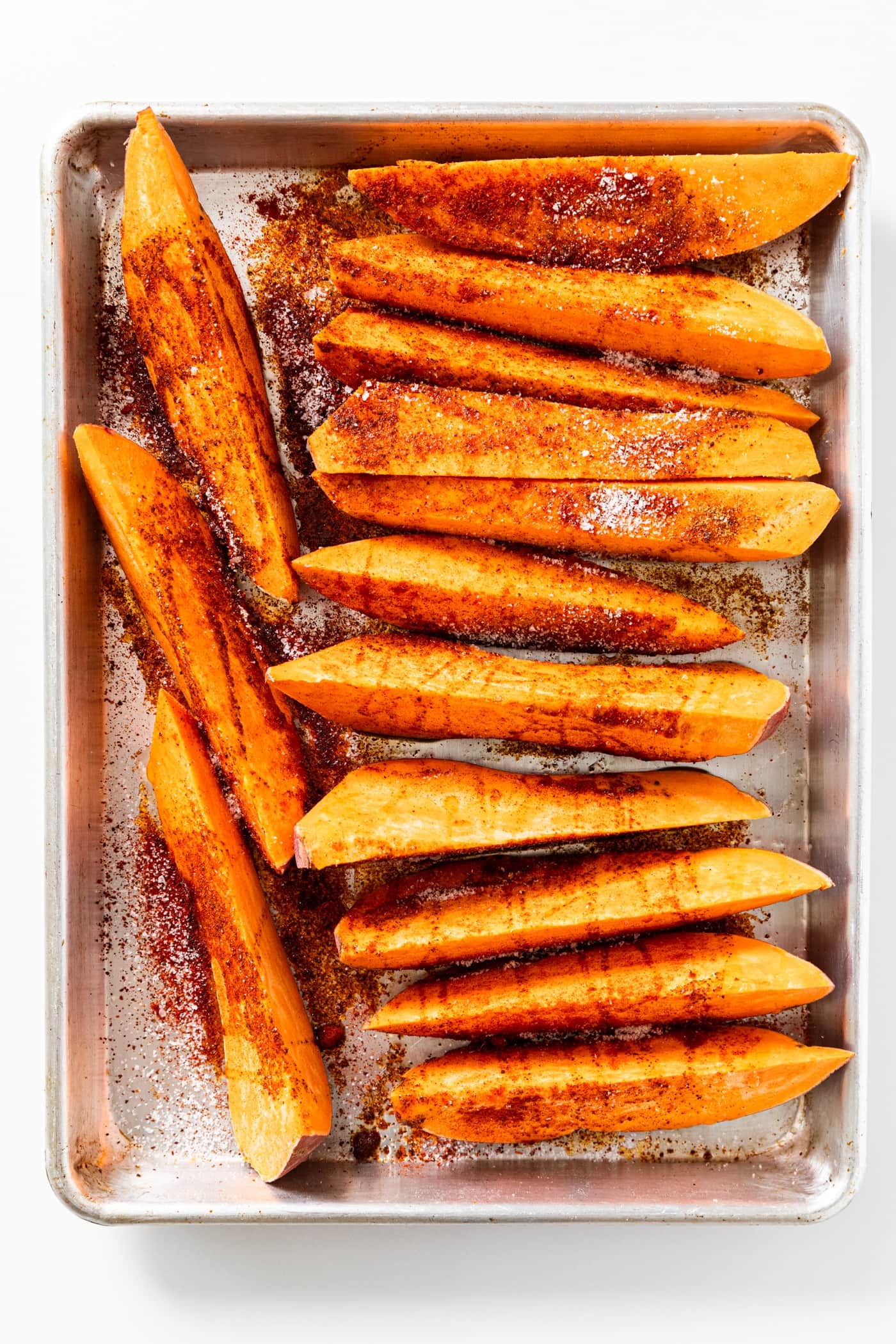 Uncooked wedges of orange colored sweet potatoes sitting on metal roasting pan all on white countertop topped with spices.