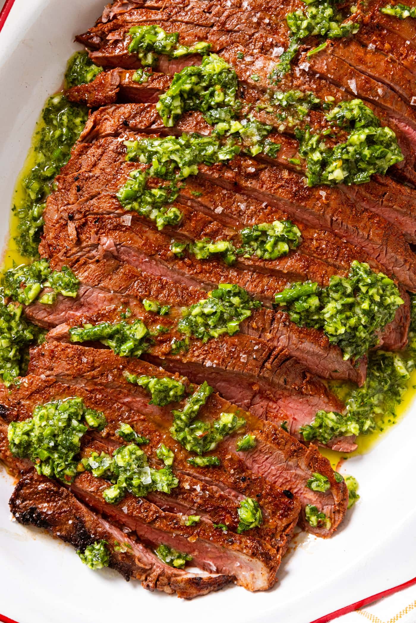 White serving platter with red rim filled with grilled flank steak topped with chimichurri sauce.