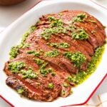 White serving platter with red rim filled with grilled flank steak with green chimichurri sauce on top with lime and orange in background.