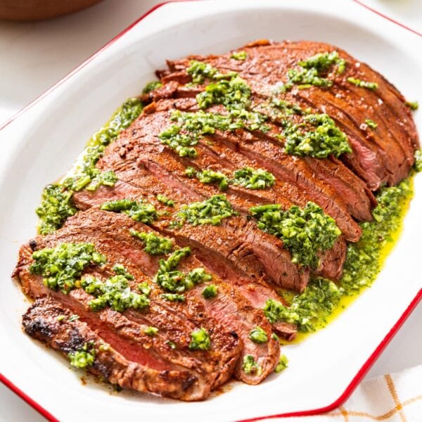 White serving platter with red rim filled with grilled flank steak with green chimichurri sauce on top with lime and orange in background.