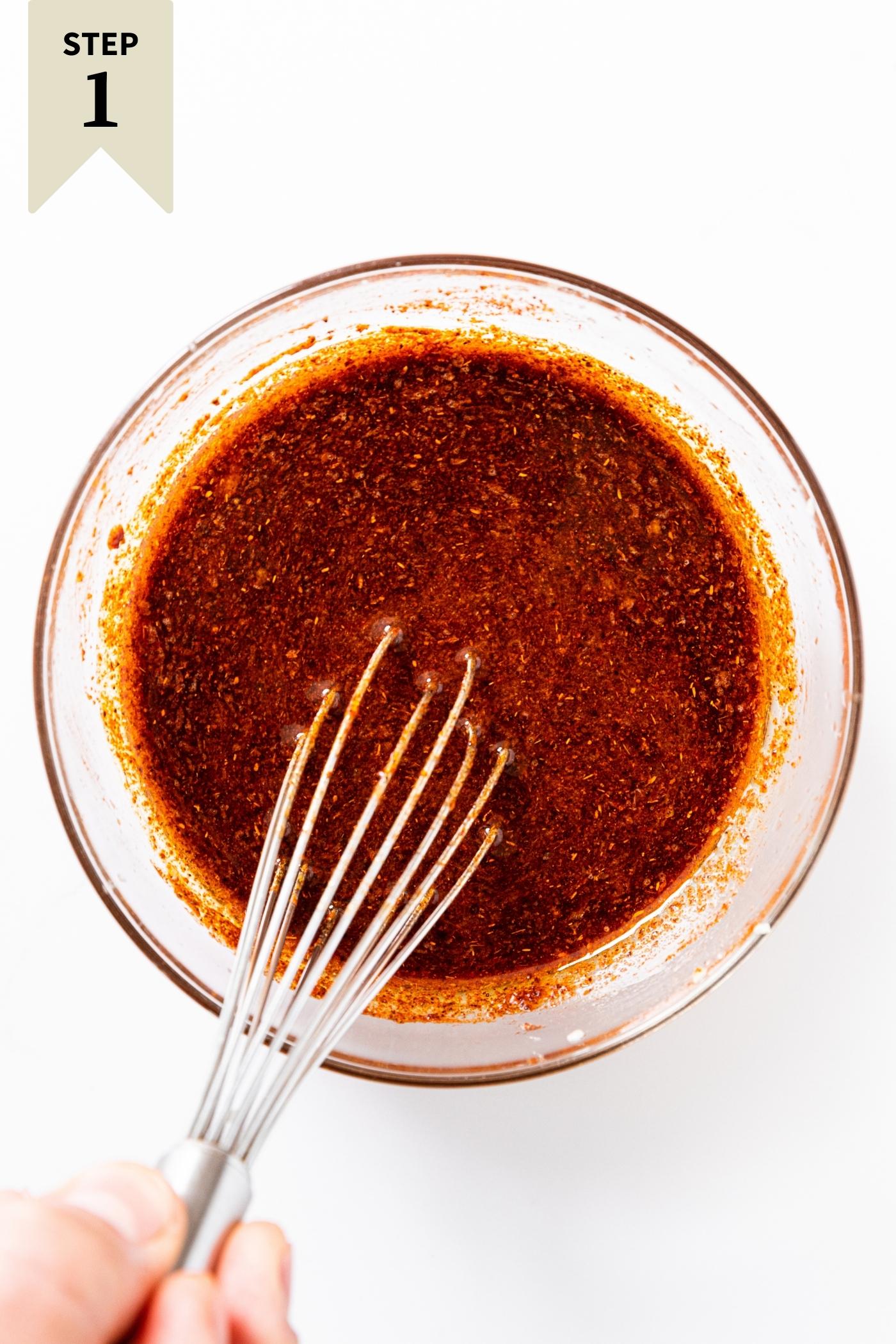White countertop with glass bowl filled with brownish red marinade inside with whisk being held by hand.