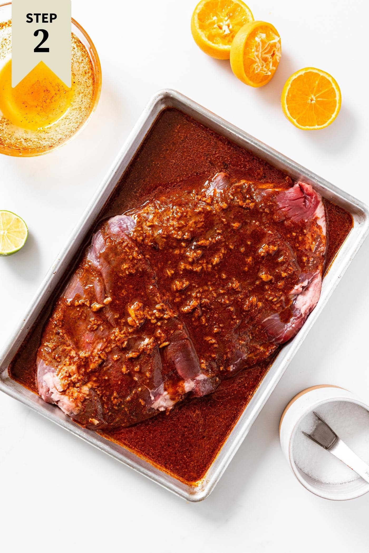 White countertop with metal pan filled with uncooked flank steak with marinade poured all over the top with used oranges and empty marinade bowl beside.