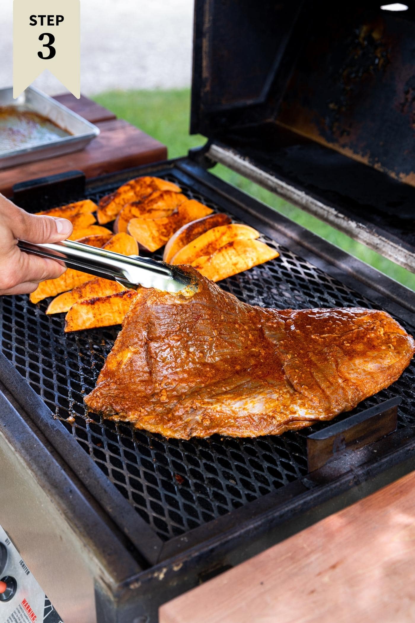 Tongs putting raw piece of flank steak on grill topped with reddish colored marinade with sweet potato wedges in background.