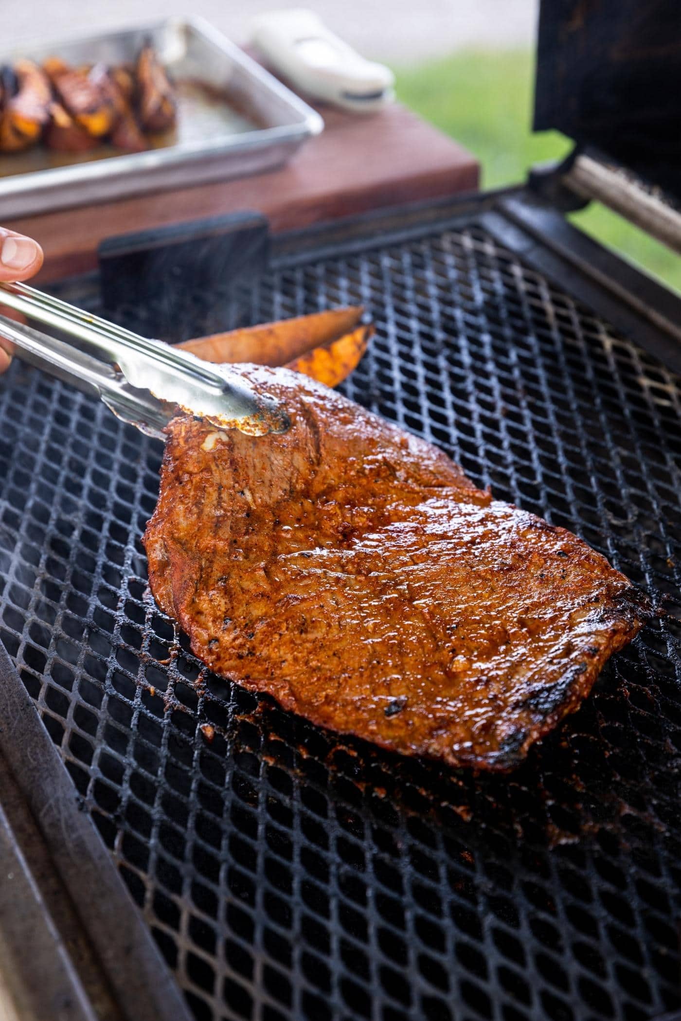 Tongs holding large piece of grilled flank steak after it grilled on the grill.