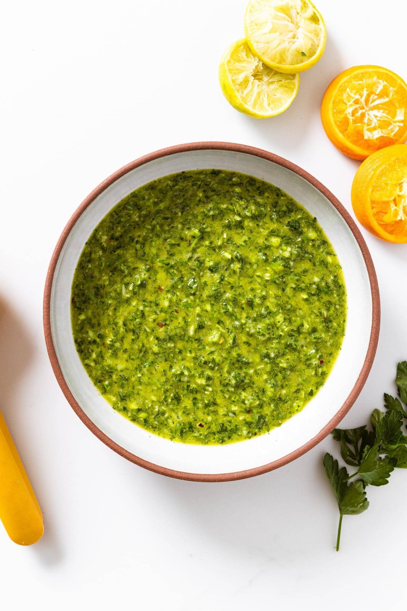 White countertop with stoneware bowl with brown rim filled with garlic scape chimichurri with used oranges and limes sitting beside.