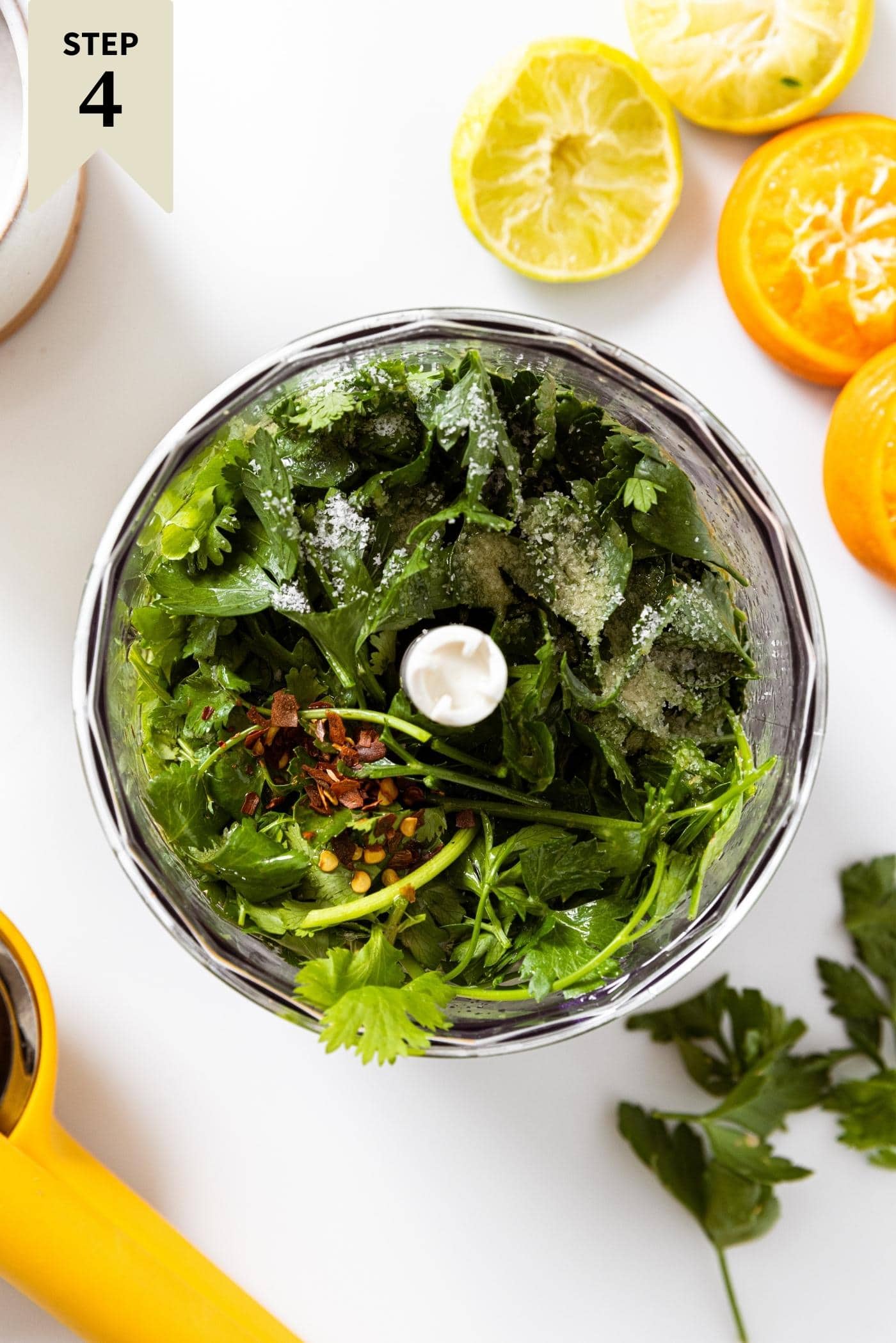 Food processor filled with parsley and cilantro with used oranges and limes sitting on white countertop.