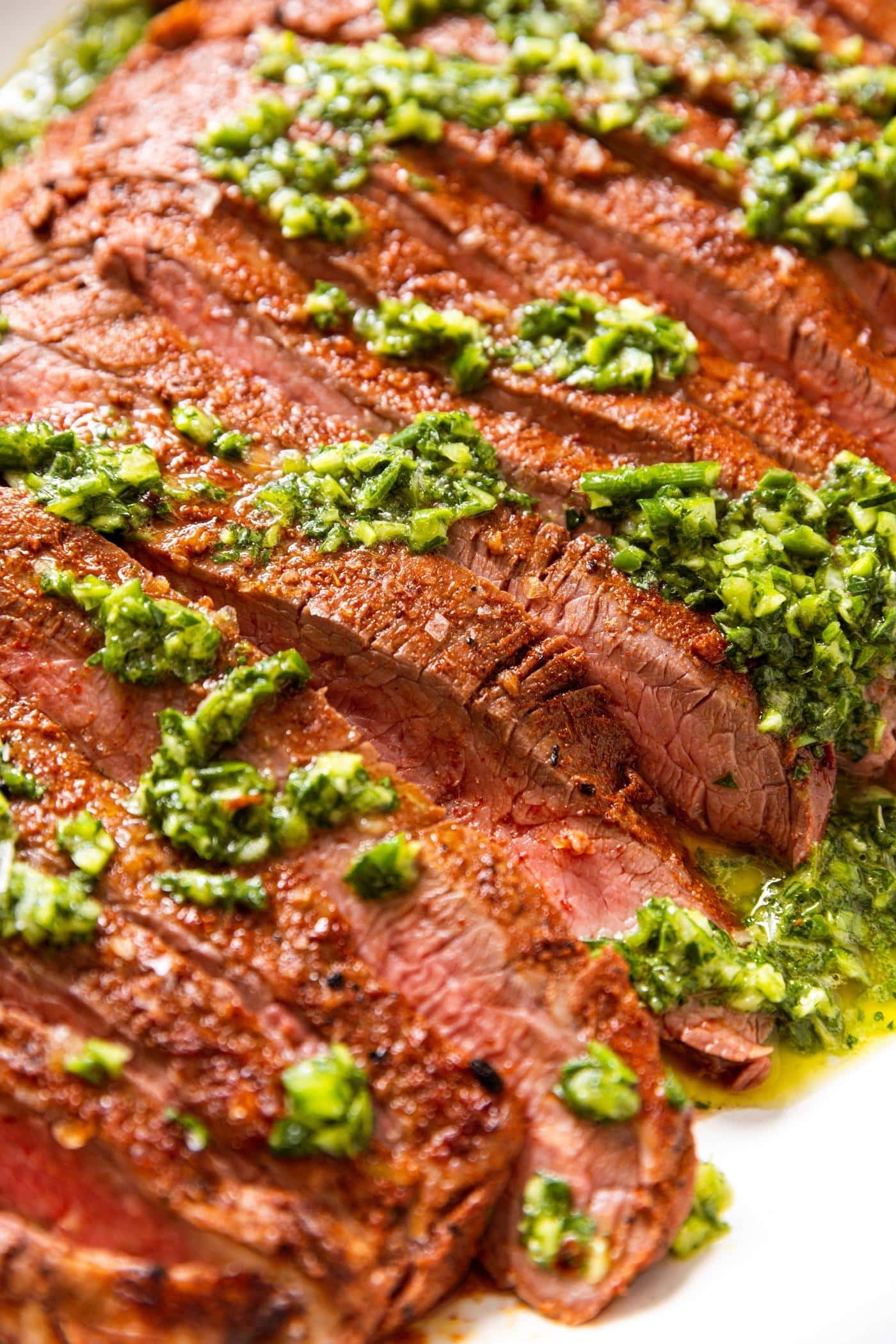 Slices of grilled flank steak sitting on white serving platter toped with green colored garlic scape chimichurri.