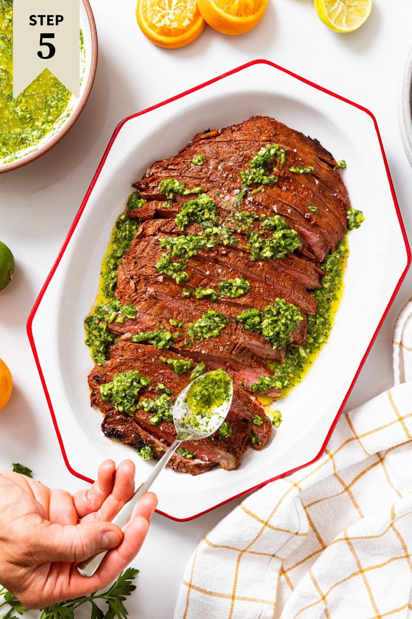 Grilled flank steak sitting in white angular serving platter with hand spooning on garlic scape chimichurri sauce.