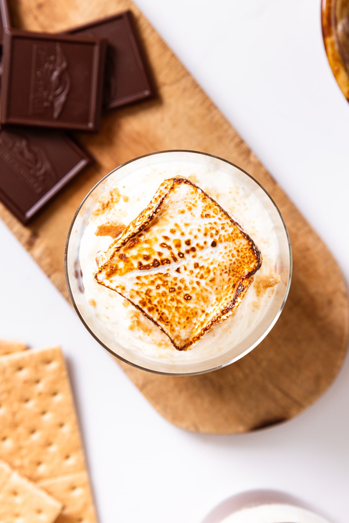 Charred marshmallow on top of whipped cream sitting in glass all on small wood cutting board with chocolate and graham crackers.