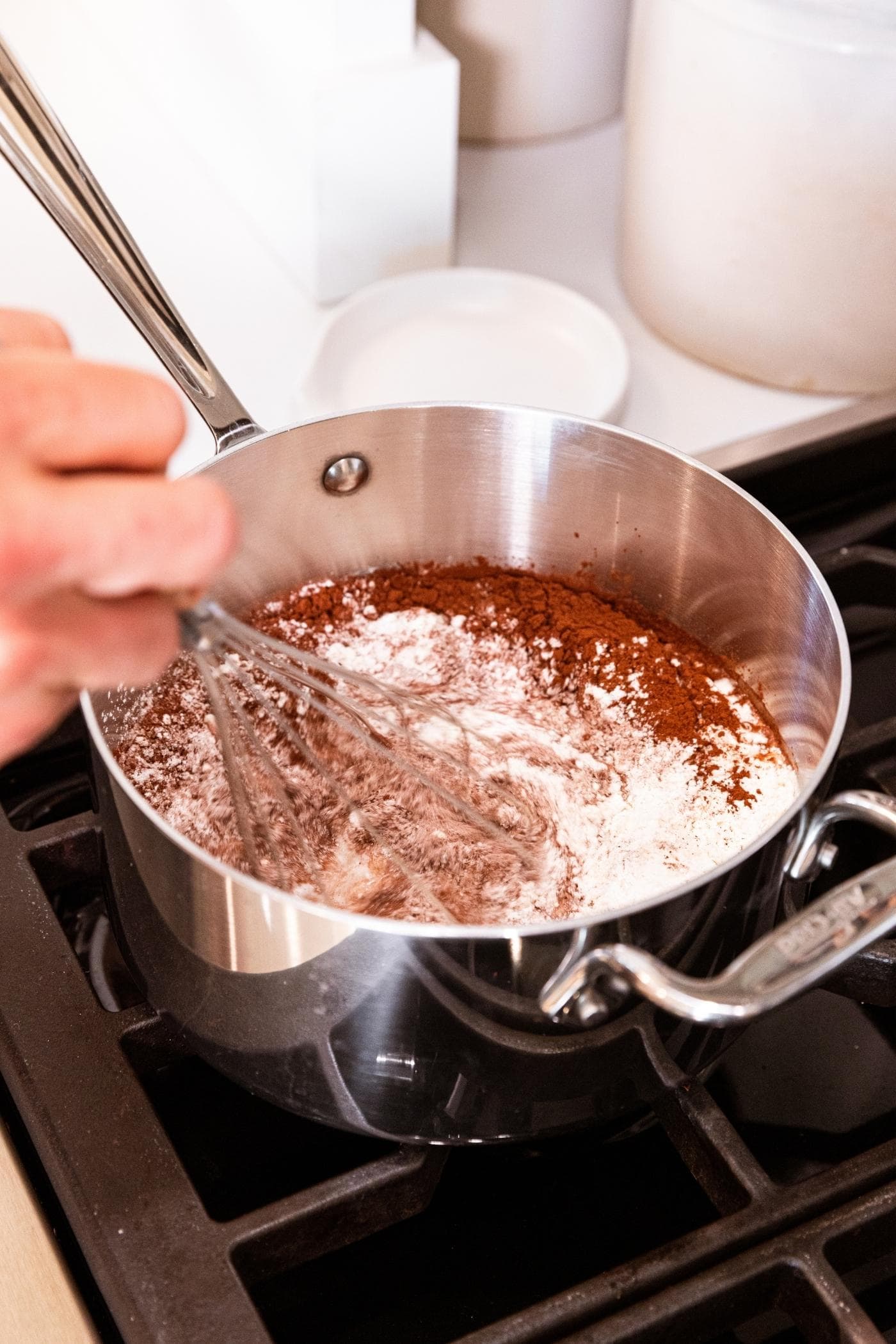 Metal saucepan sitting on stovetop with hand holding whisk whisking ingredients in pan including flour, milk, and cocoa.