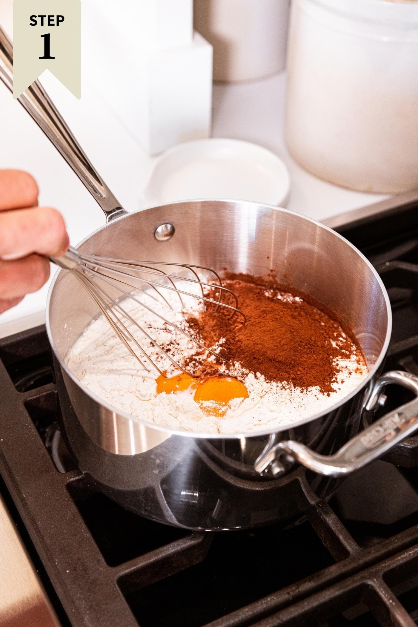Metal saucepan sitting on stovetop filled with eggs, cocoa, milk, flour, and sugar with hand holding whisk.