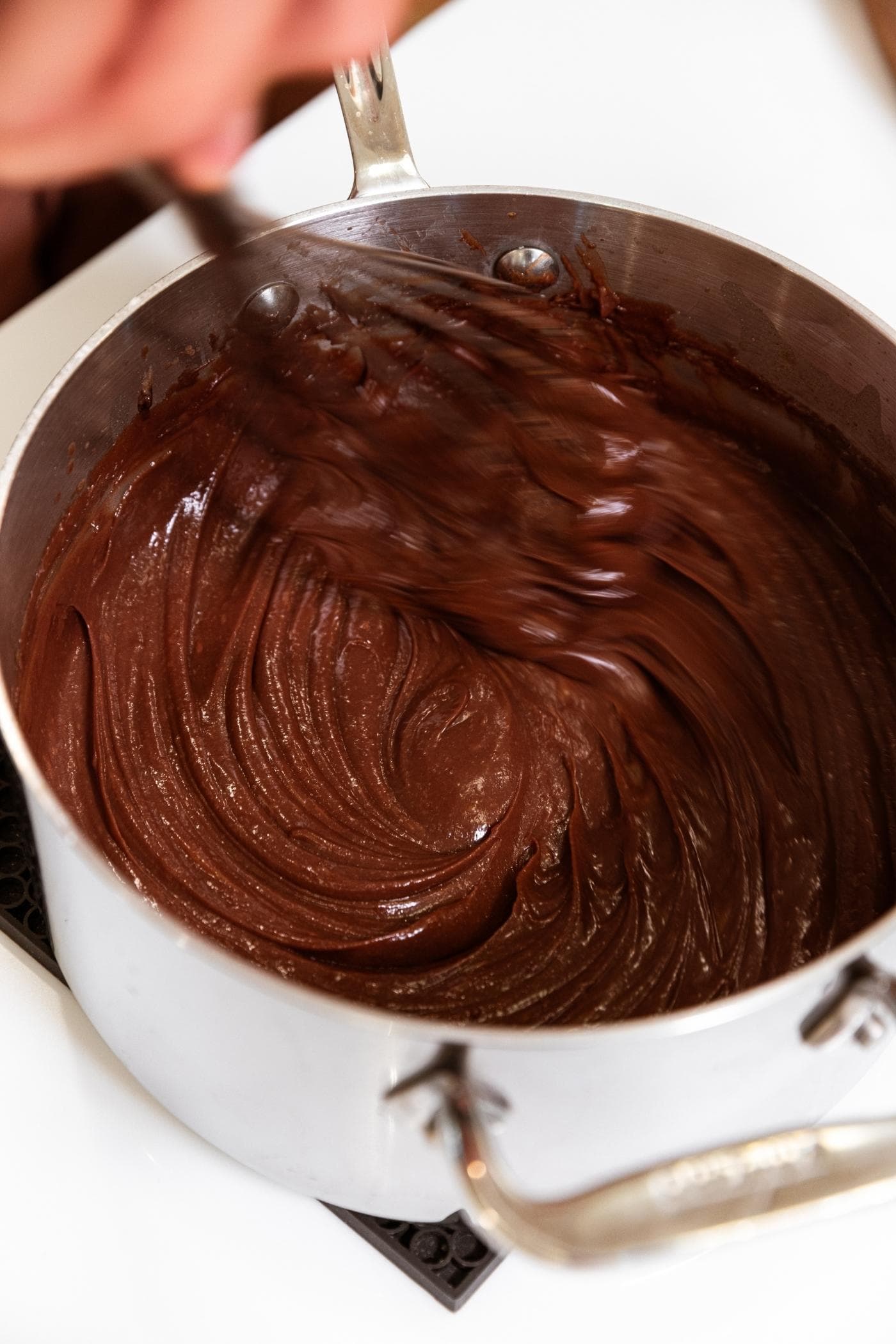 Chocolate mixture in metal saucepan sitting on white countertop with hand whisking mixture together.