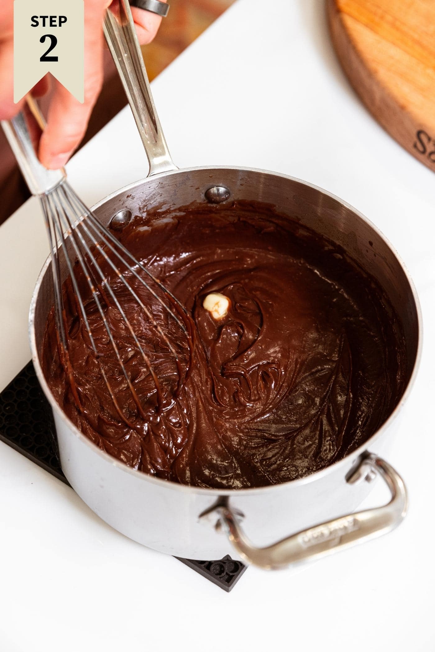 Metal saucepan sitting on white countertop with piece of butter and vanilla extract in pan on top of chocolate mixture with hand holding whisk.