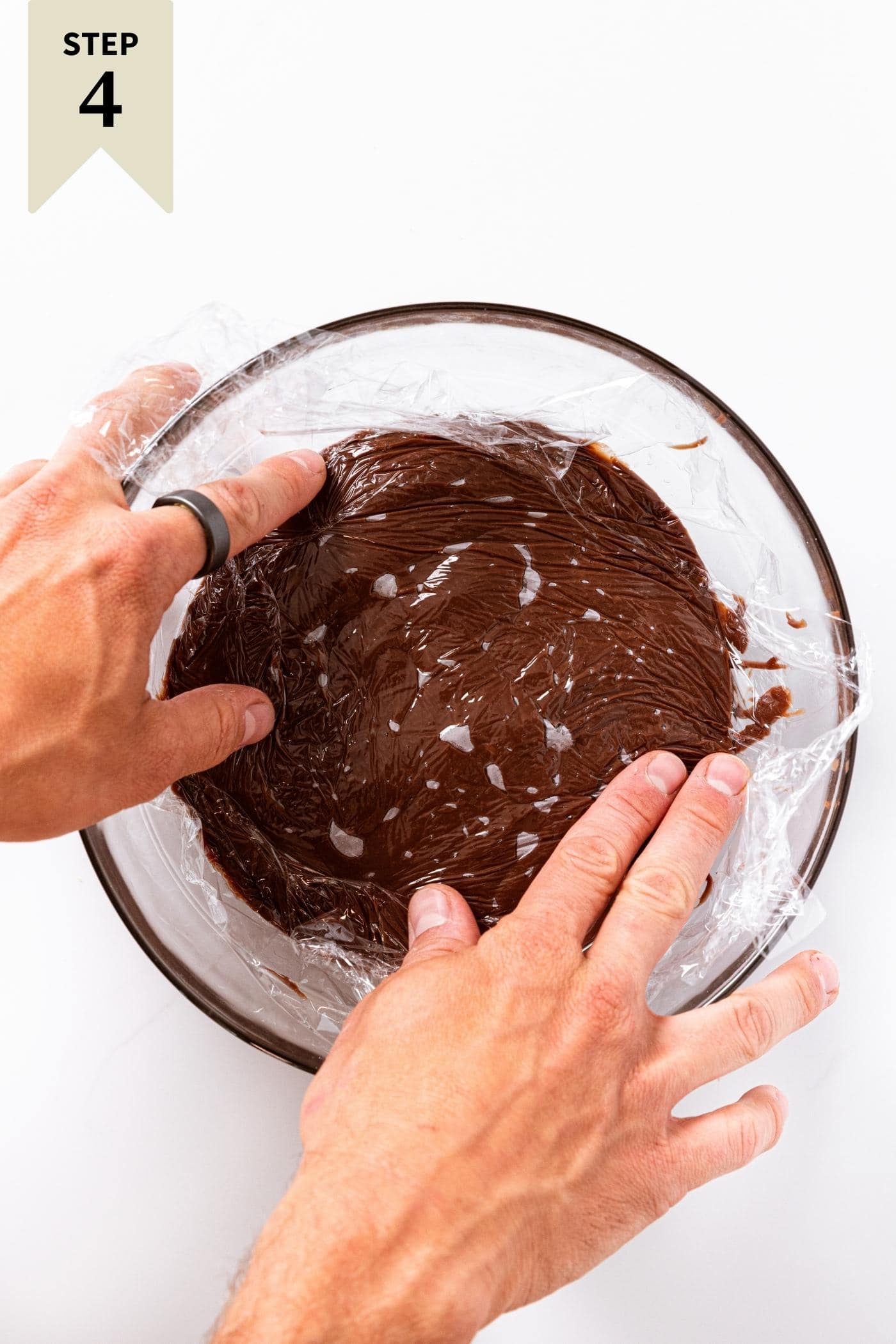 White countertop with glass bowl filled with chocolate pudding with two hands pressing plastic wrap directly on surface of pudding.