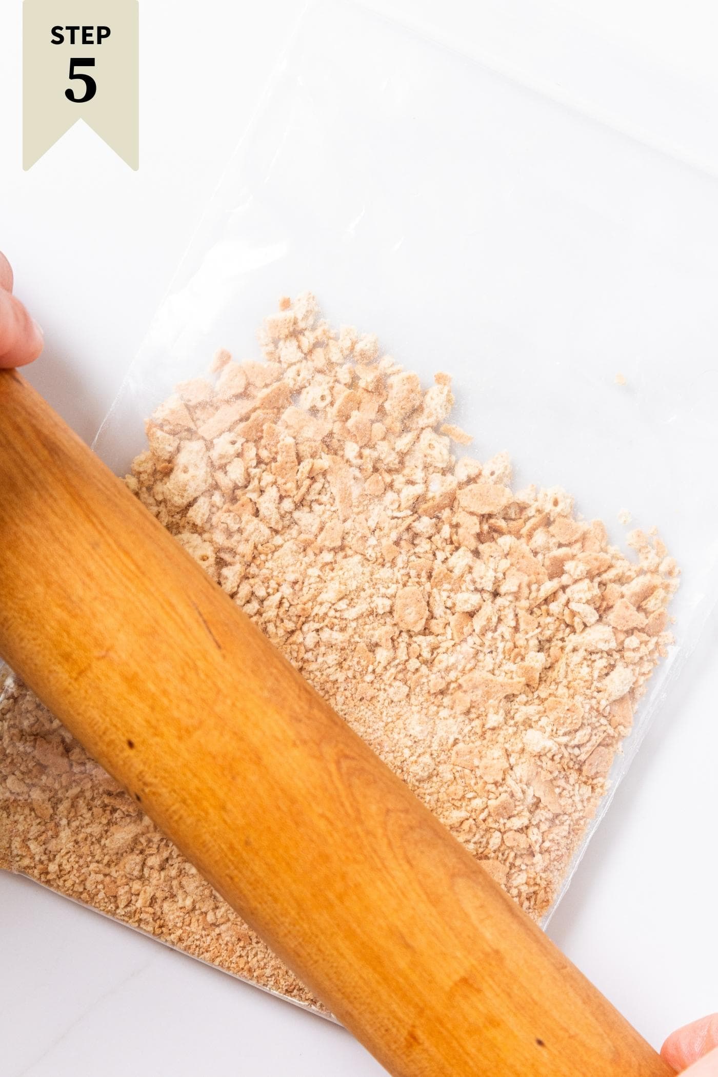 White countertop with plastic bag with graham cracker crumbs inside with rolling pin crushing more crumbs.
