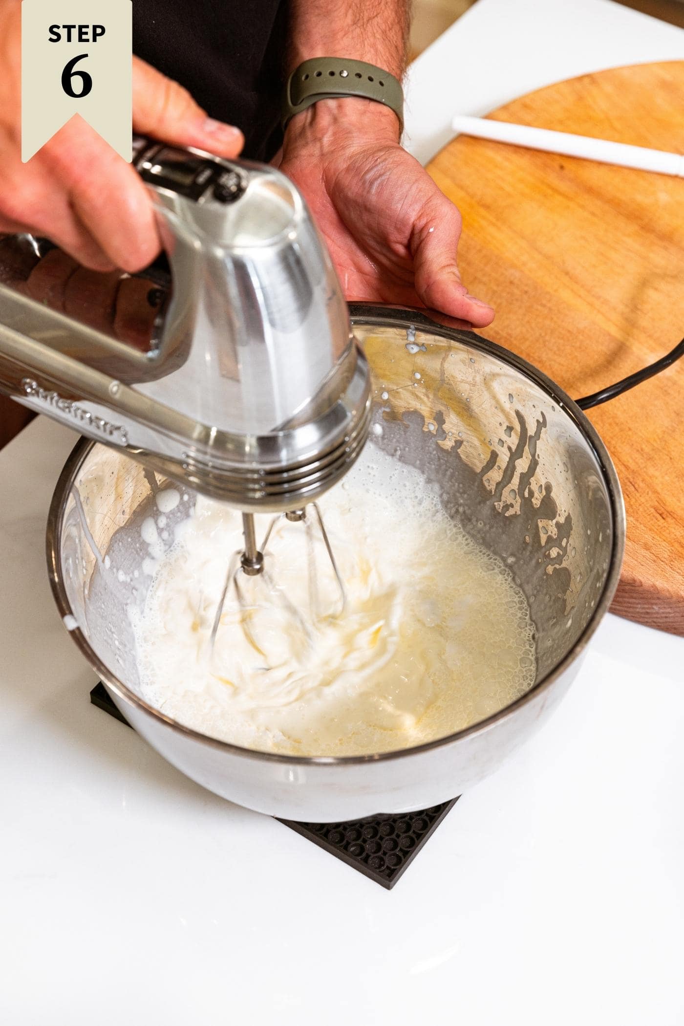 Metal bowl filled with cream being mixed by hand mixer all on white surface.