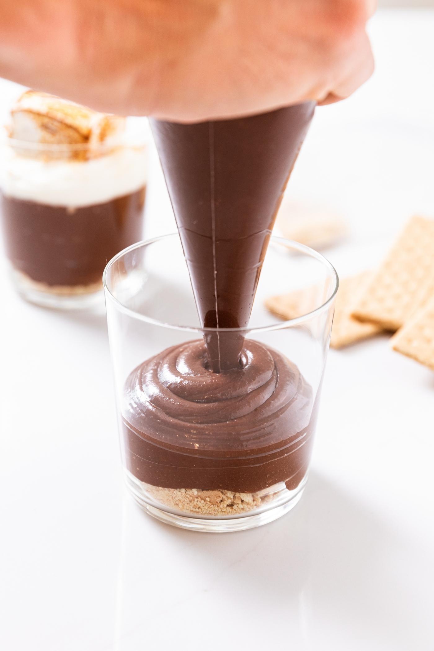 Hand holding piping bag filled with chocolate pudding filling up a clear glass with graham cracker crumbs along base.