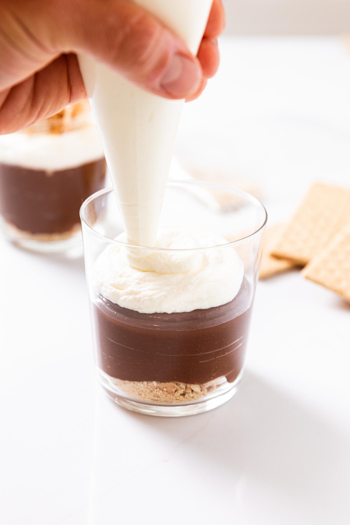 Clear glass container being topped with whipped cream from piping bag held by a hand all on white countertop.
