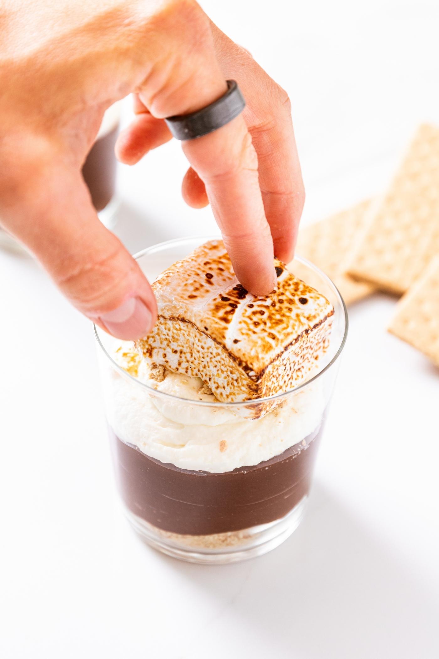 Hand placing charred square marshmallow on top of whipped cream in pudding cup sitting on white countertop.