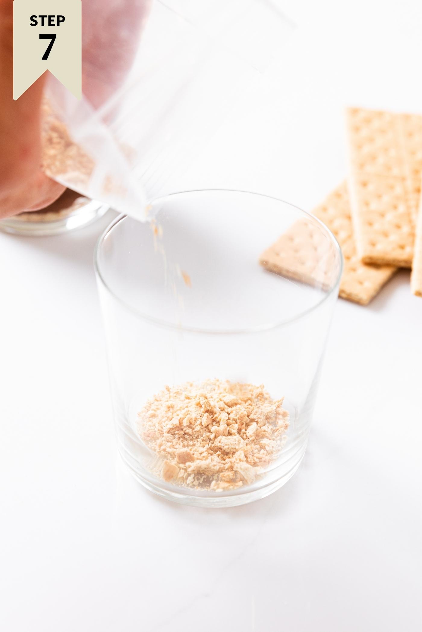 Clear glass sitting on white surface being filled from plastic bag with graham cracker crumb base.