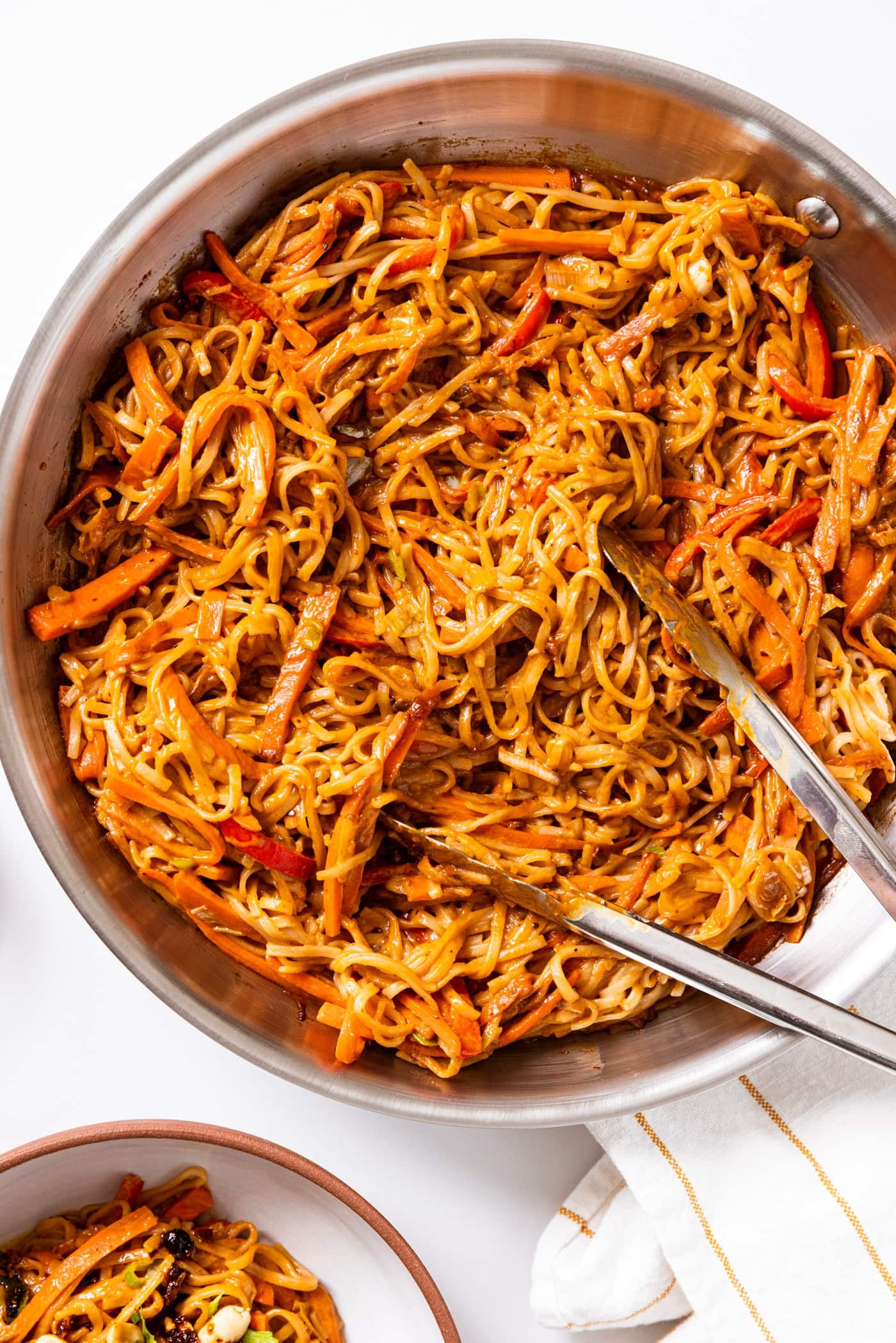 Large skillet filled with spicy peanut noodles sitting on white countertop with tongs inside.
