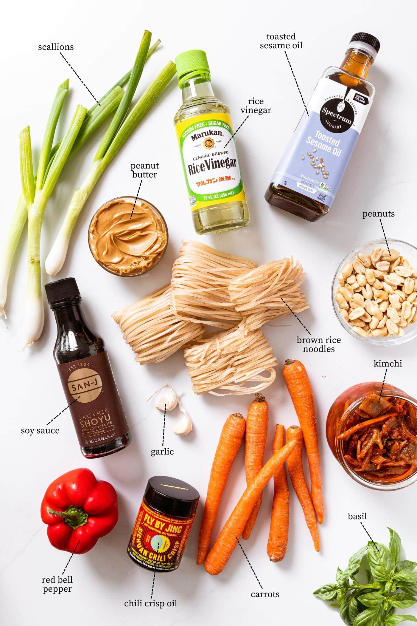 White countertop with lots of ingredients needed to make spicy peanut noodles including carrots, pepper, scallion, peanuts, kimchi, and more.