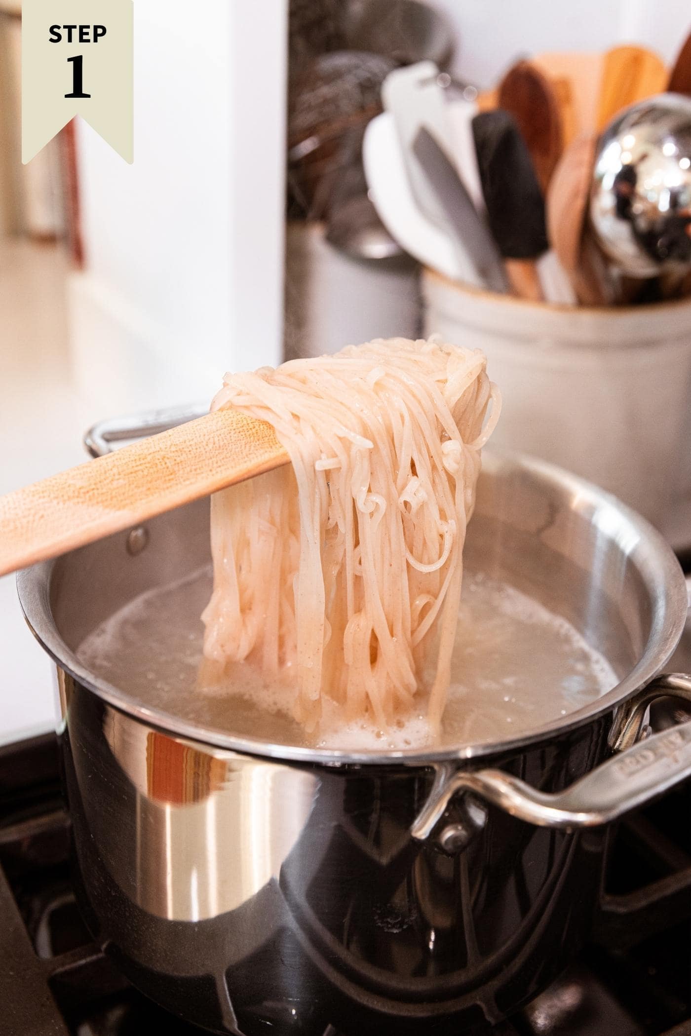 Wooden spatula pulling noodles from large metal stockpot sitting on stovetop.