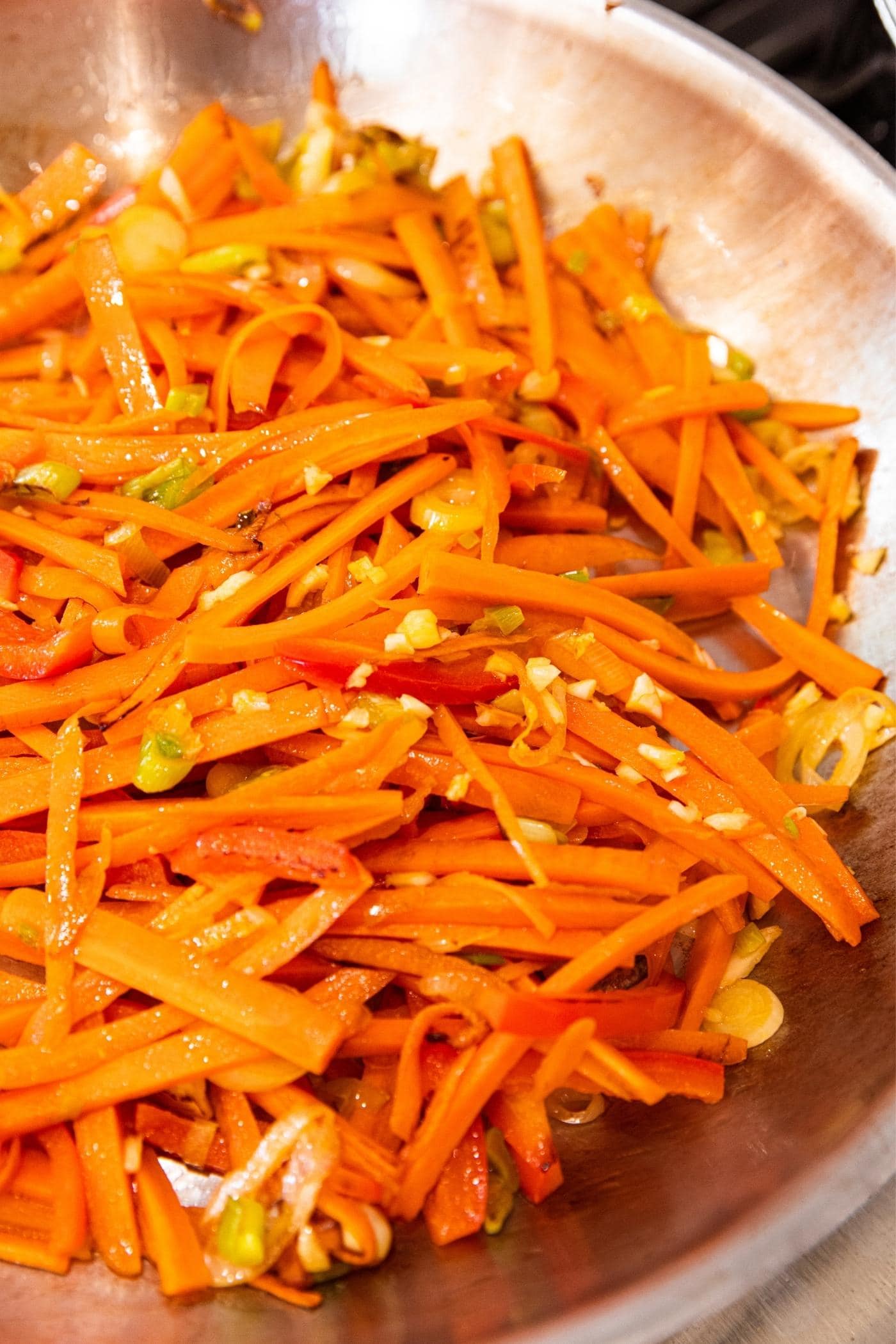 Sticks of carrots and red bell peppers sitting in metal skillet on stovetop with small bits of garlic chopped up.