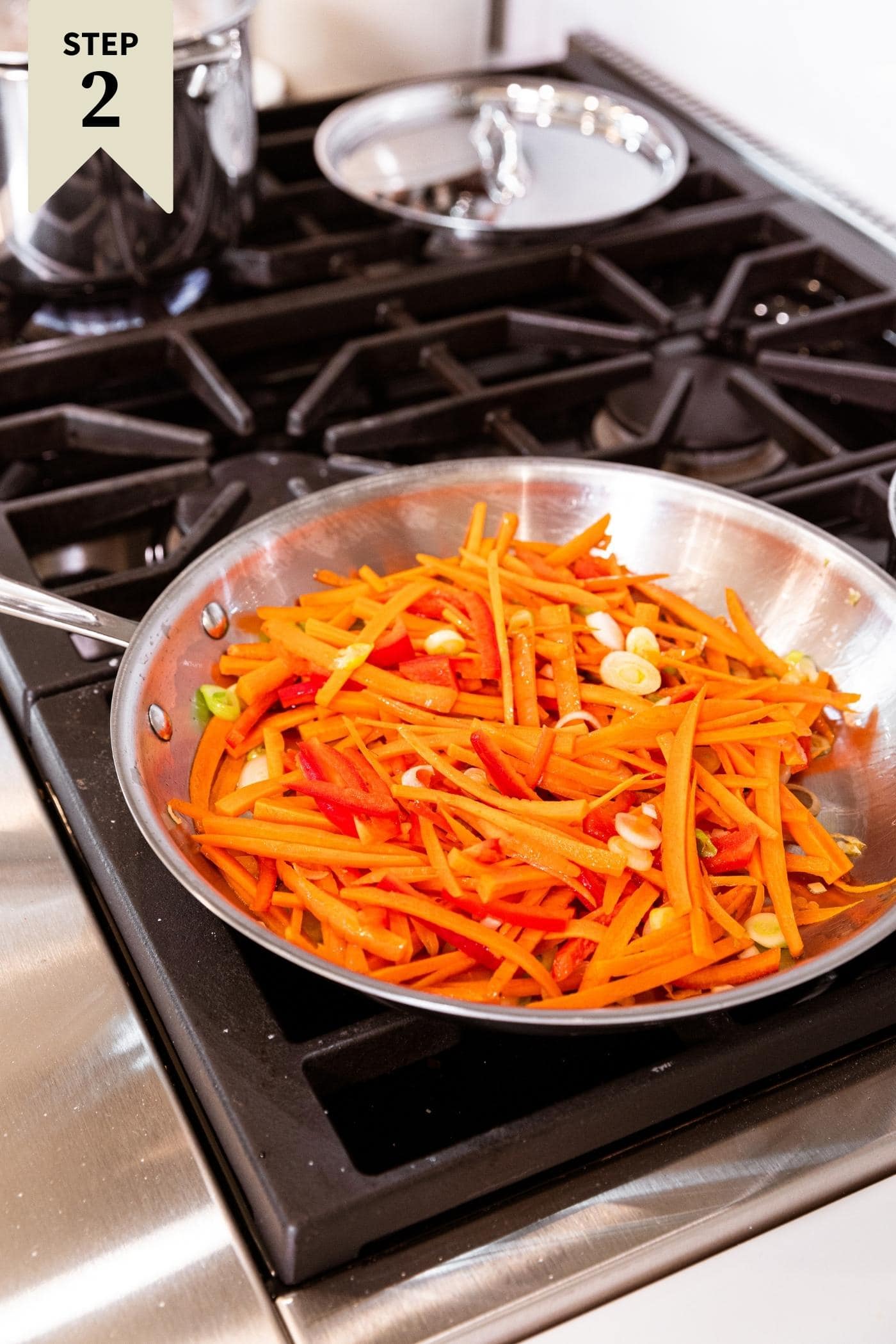 Large metal skillet sitting on stovetop filled with sticks of carrots, red bell peppers, and scallions.