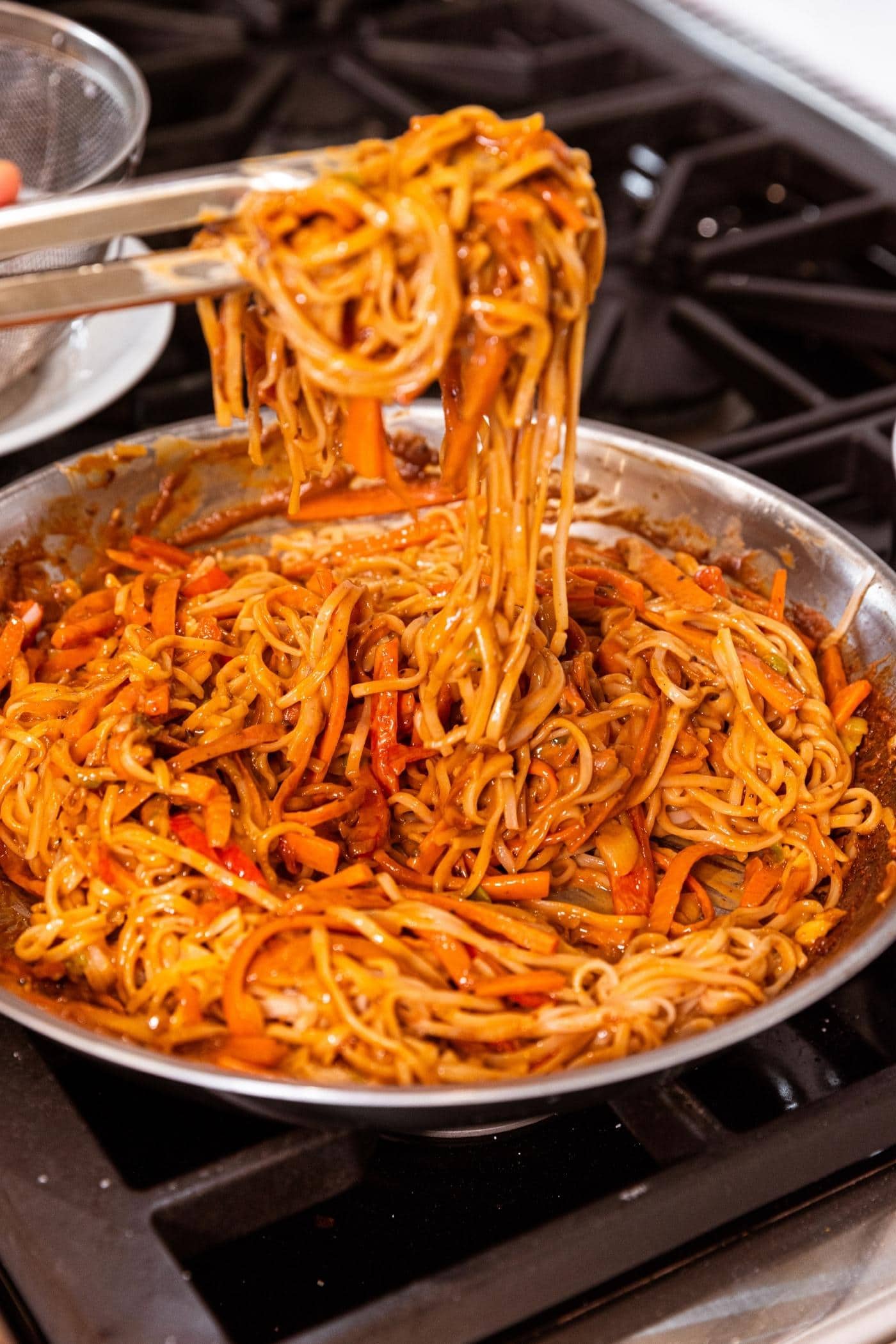 Tongs mixing rice noodles into peanut sauce and vegetables in metal skillet.