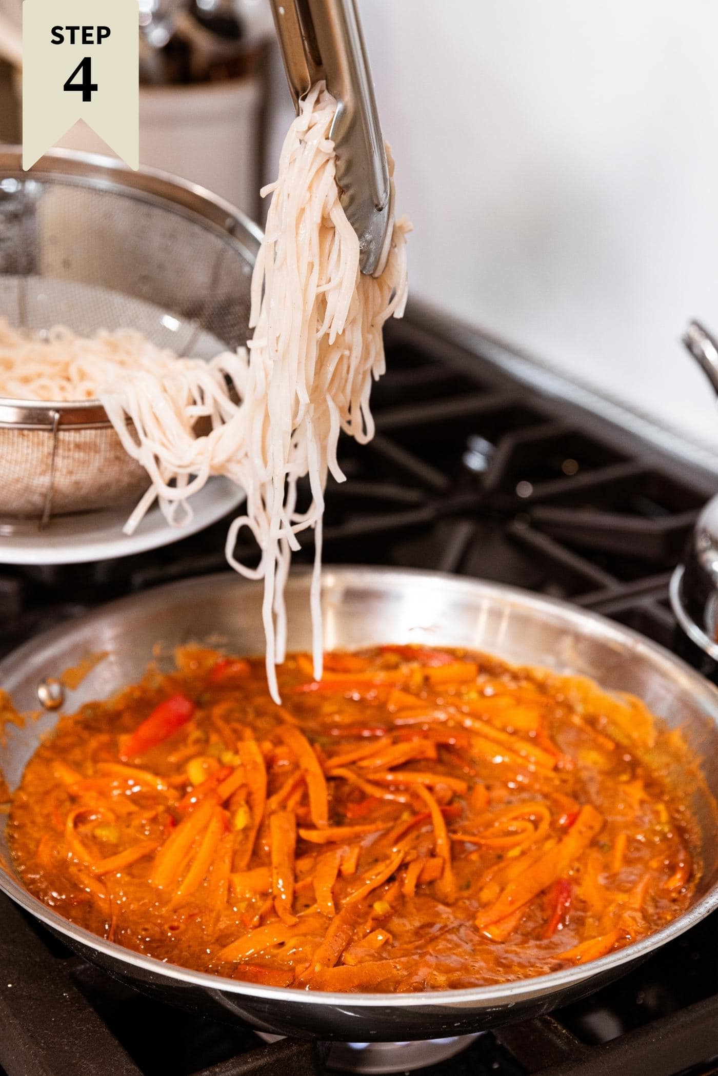 Tongs holding cooked rice noodles dangling them over peanut sauce with vegetables in skillet below all on stovetop.
