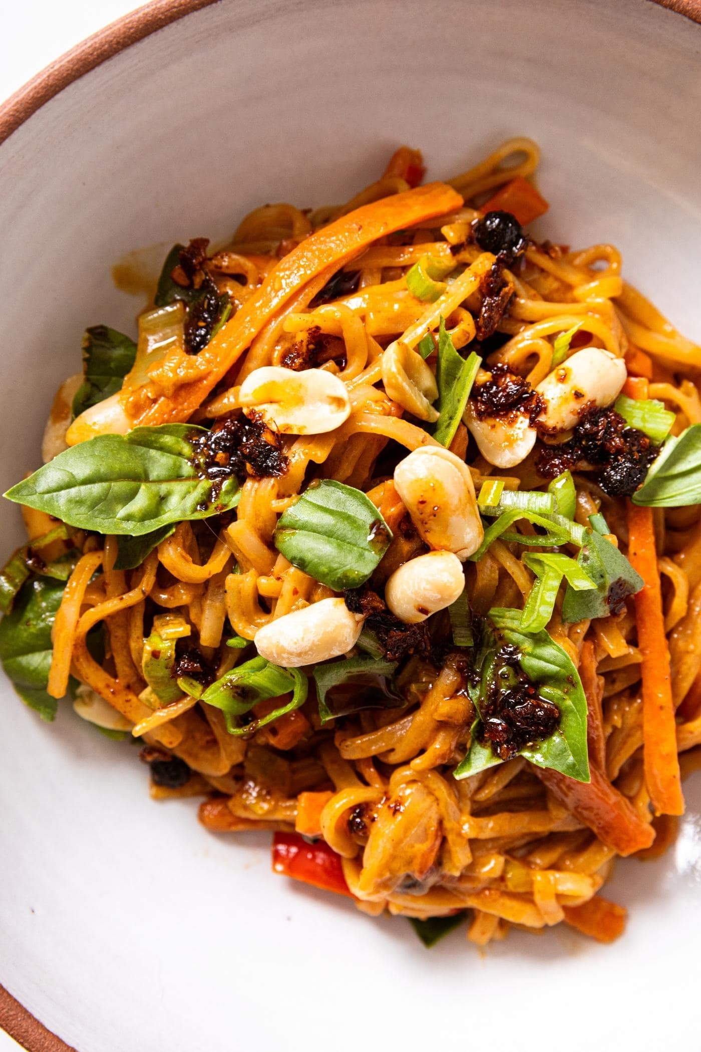 White stoneware bowl with brown rim filled with spicy peanut noodles topped with leaves of basil, peanuts, and chili sauce.