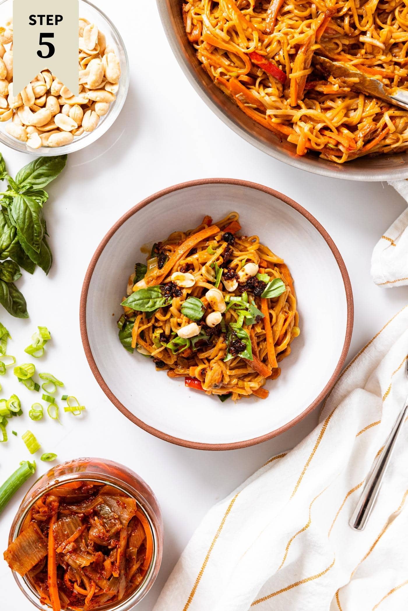 White bowl with brown rimming sitting on white countertop filled with spicy peanut noodles garnished with basil leaves and peanuts with other parts of the meal sitting around.